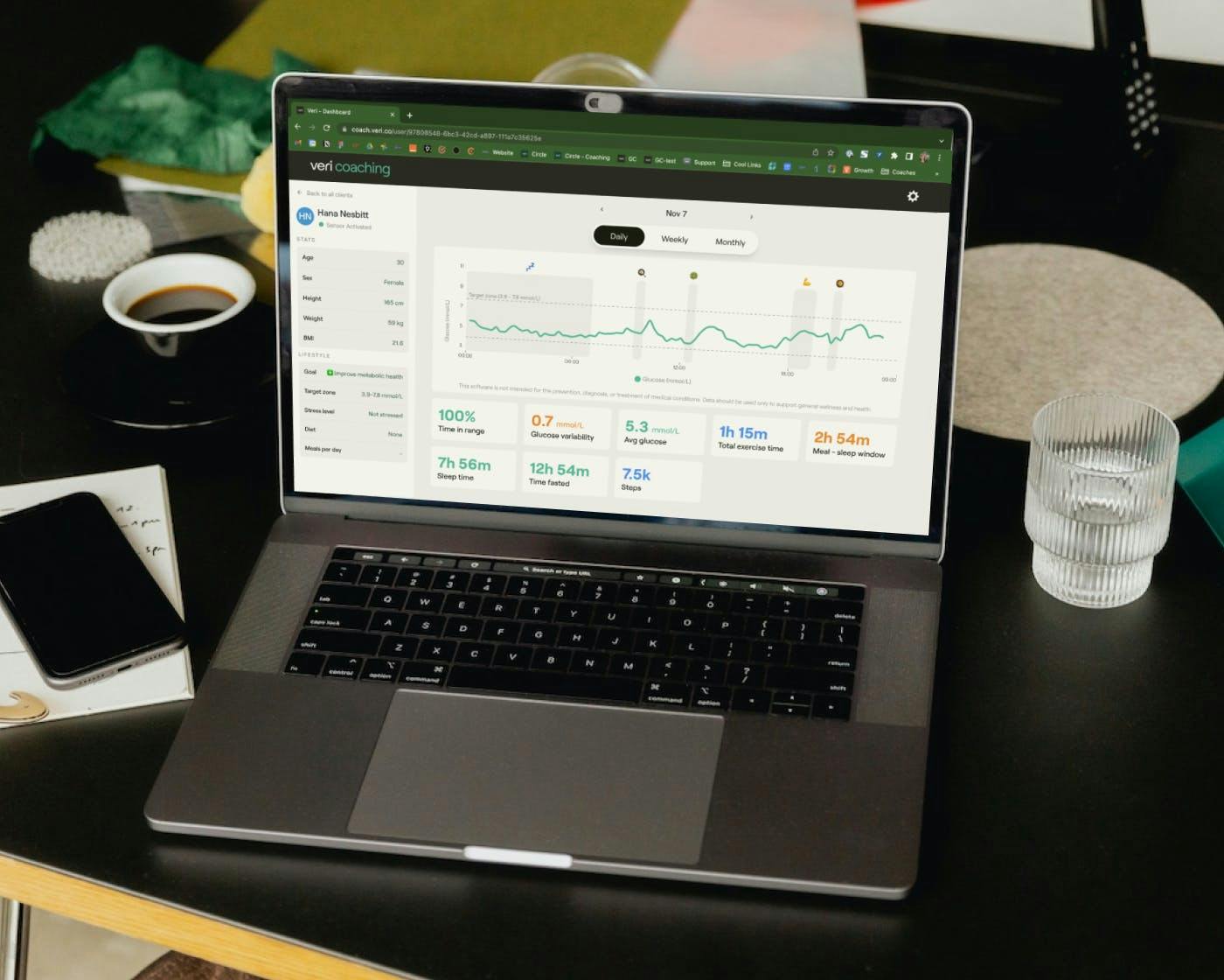 A laptop sits on a coach's desk, with the coaching web dashboard open on it, display a client's metabolic health data. 