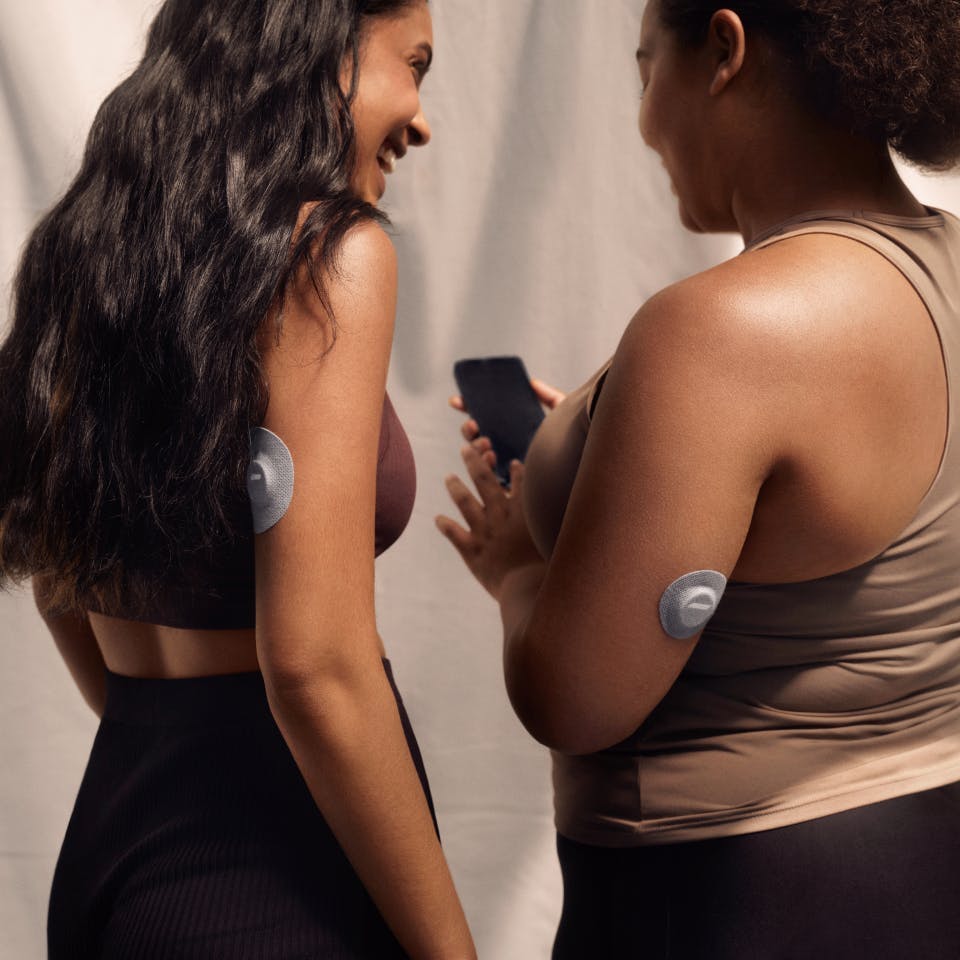Two women wearing CGMs (continuous glucose monitors) share a glance at the Veri app on one woman's phone