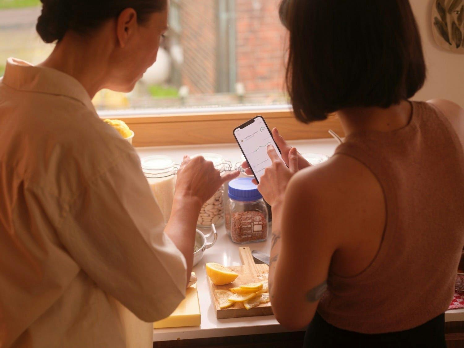 two women standing over a kitchen counter and studying data on a phone screen
