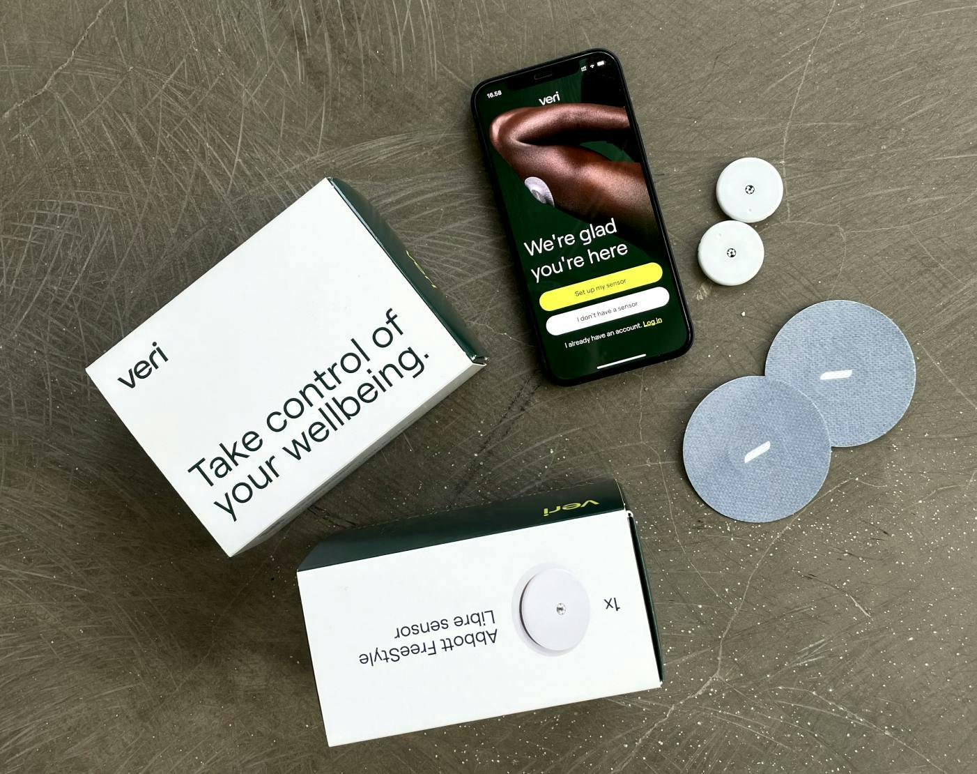A Veri box is laid out on a table next to two CGM continuous glucose monitors and a mobile phone with the Veri app