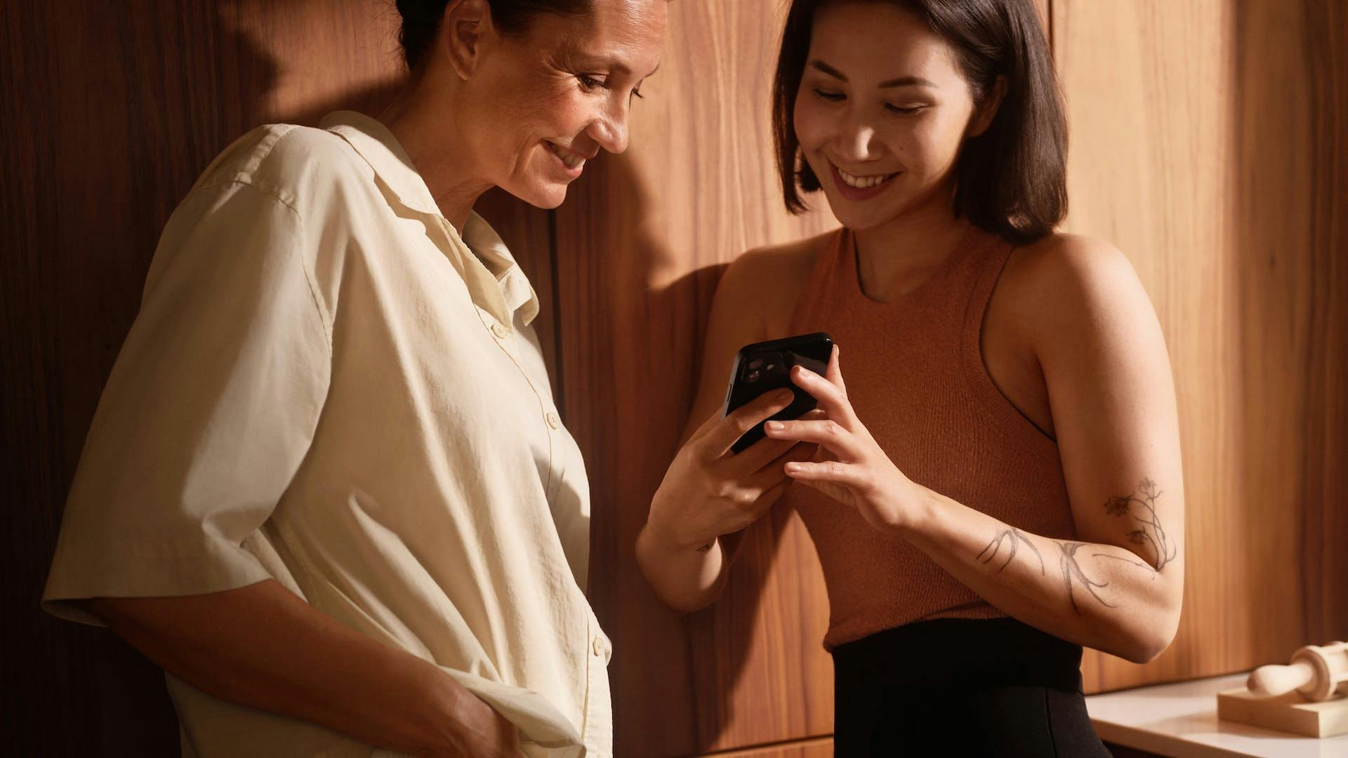 two women standing side by side and smiling while looking at a phone screen